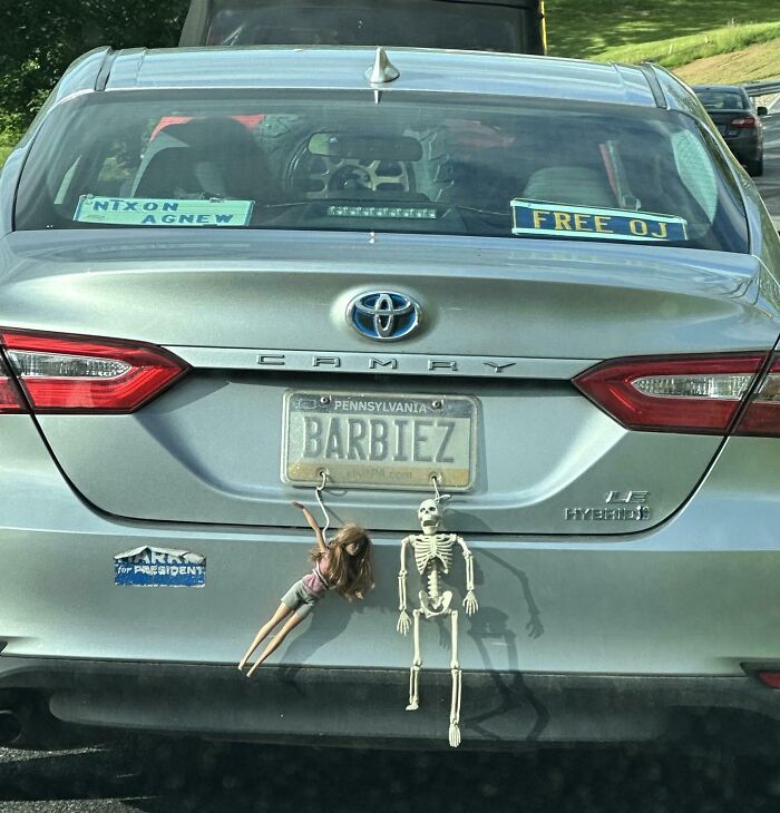 Silver Toyota Camry with funny bumper stickers and hanging Barbie doll and skeleton decorations on the rear of the car.