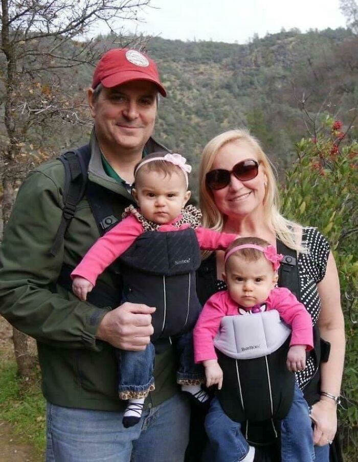 Pareja con dos bebés en mochilas porta bebés mostrando expresiones incómodas en foto familiar icónica.