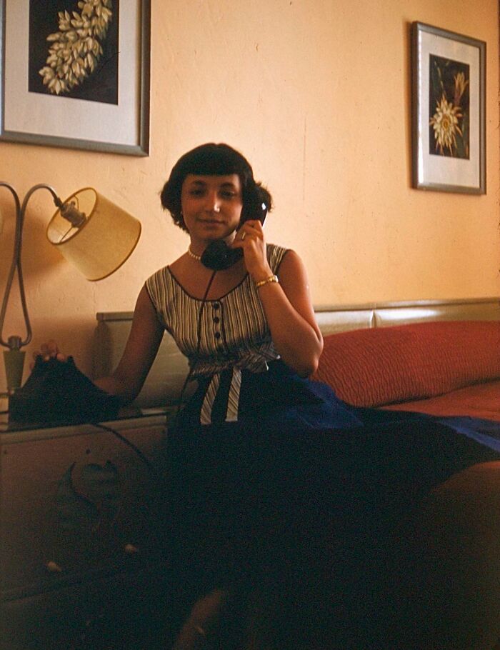 Woman in vintage dress sitting on hotel bed, talking on a rotary phone, vintage vacation pic from before cell phone cameras.