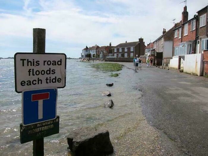 Road sign warning this road floods each tide, confusing drivers by water covering the street near residential houses.