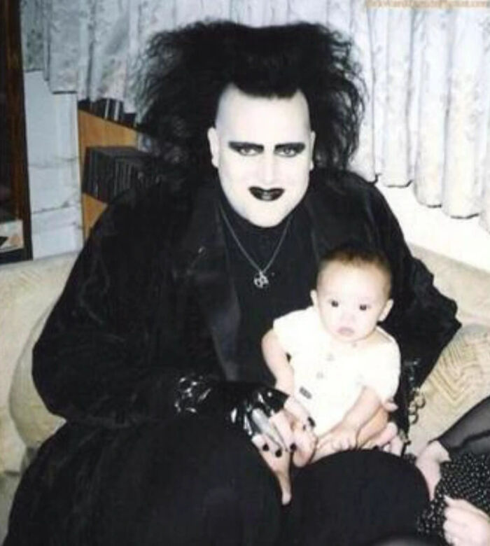 Man in gothic makeup and outfit holding baby in a family photo that later became laughed or cringed at online.