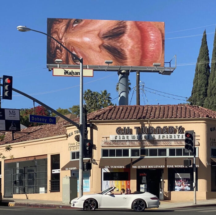 Funny and weird billboard with a giant close-up face over a street intersection and a convertible car below.