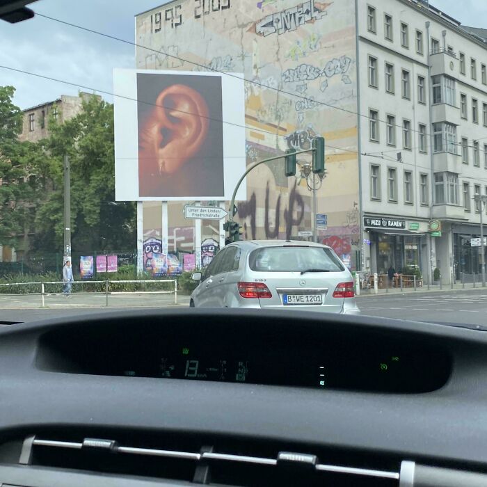 Weird billboard displaying a large close-up ear image on a city street with cars and graffiti in the background.