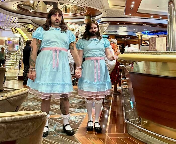 Two men dressed in creative Halloween costumes wearing matching blue dresses with pink ribbons inside a cruise ship lounge.