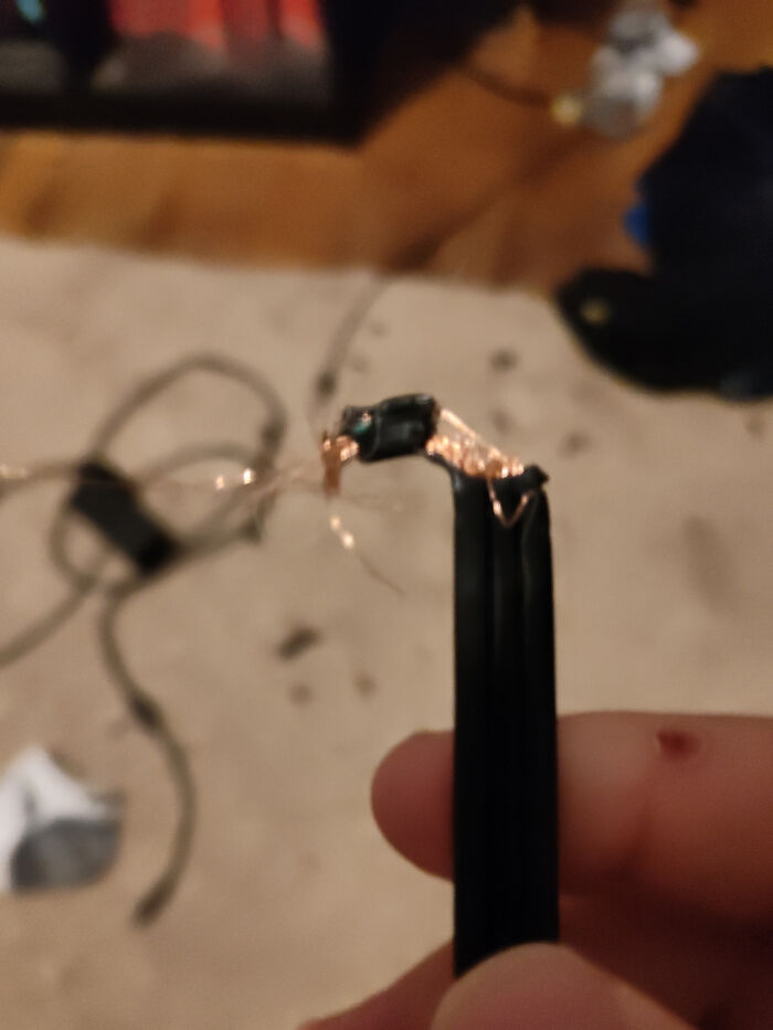 Close-up of a damaged electronic cable with exposed wires held by a person's fingers in a blurry indoor setting.