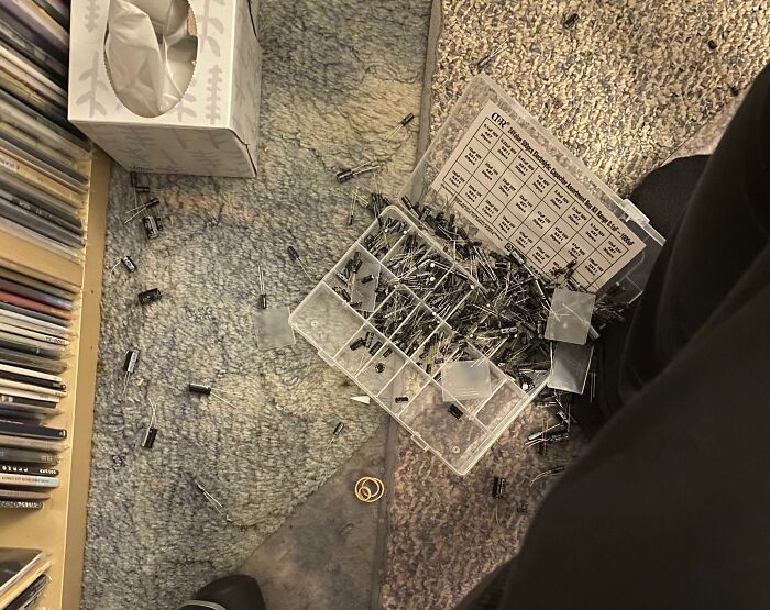 Box of electronic components spilled on carpet with scattered capacitors and resistors, illustrating mishandling of electronics and soldering.
