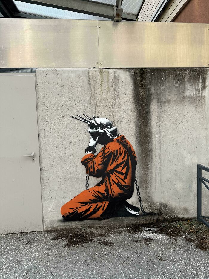 Powerful street art of a person in an orange jumpsuit, wearing a crown of thorns and shackled, painted on a concrete wall.