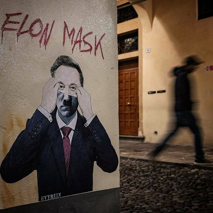 Street art depicting a man removing a mask with “Elon Mask” text on a wall in an urban setting with a blurred pedestrian.