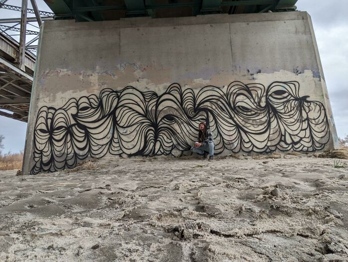 Abstract black line street art on concrete wall under bridge with person posing nearby, showcasing beautiful powerful street art.