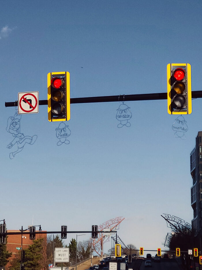 Wire-frame street art installations hanging from traffic lights above city intersection under clear sky.