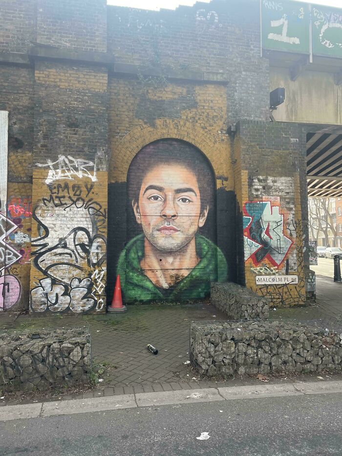Realistic street art mural of a young man with short hair and a green hoodie on a brick wall with surrounding graffiti.