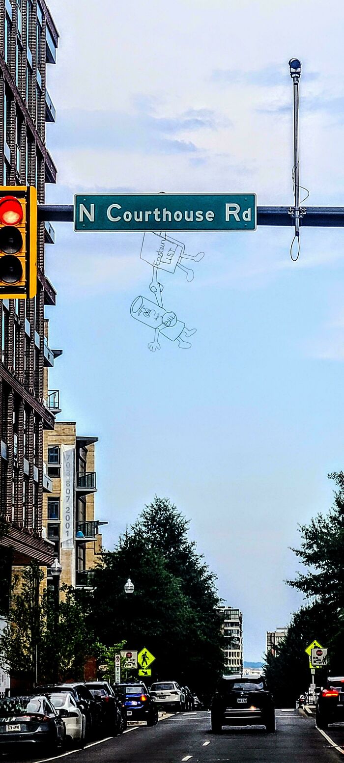 Street art of two wire-frame characters hanging from a N Courthouse Rd sign on an urban street with traffic and buildings.