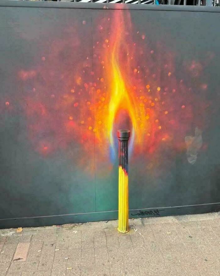 Street art featuring a painted matchstick blending into a real post with vibrant flame and glowing embers effect.