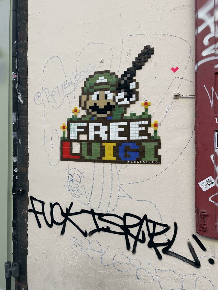 Mosaic street art of pixelated character holding a gun above colorful "FREE LUIGI" text on an urban wall with graffiti tags.