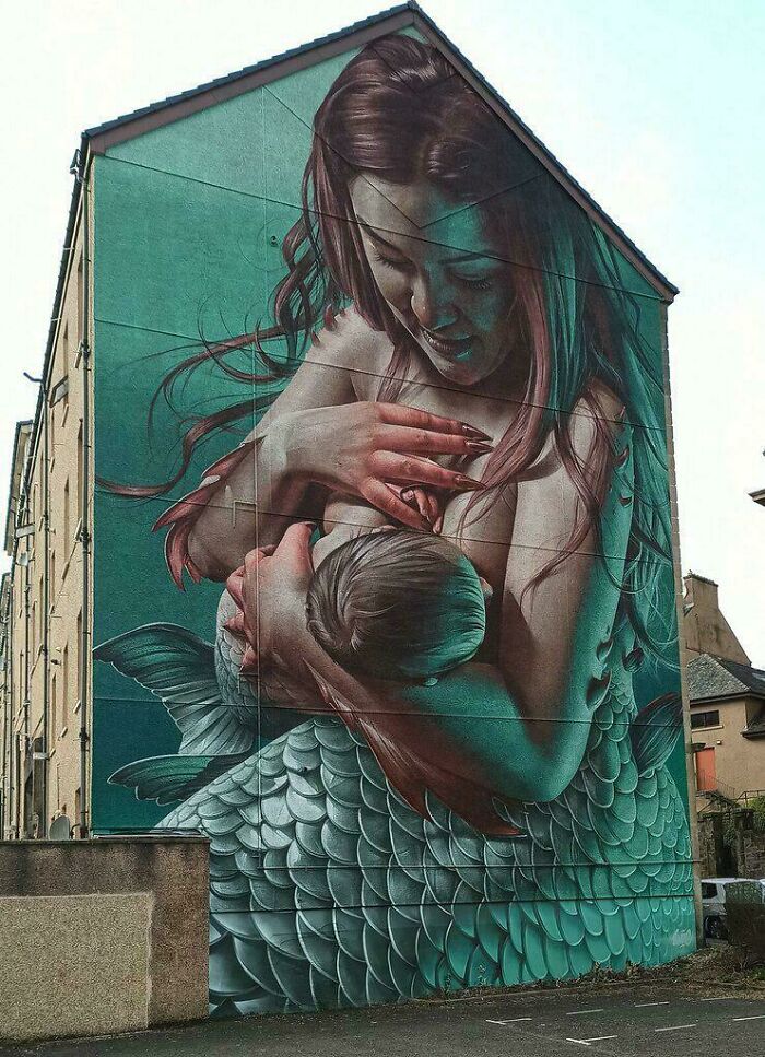Large street art mural of a mermaid mother breastfeeding her child painted on the side of a building.