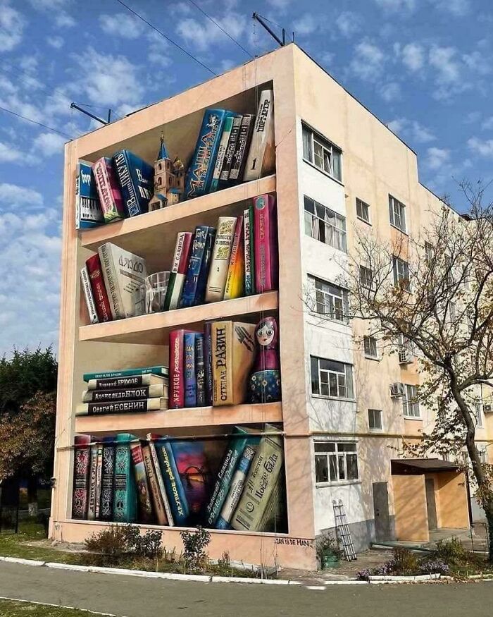 Large building painted with beautiful and powerful street art showing a bookshelf filled with colorful books and objects.