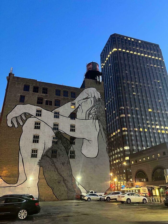 Large powerful street art mural of a seated figure on a city building wall at dusk among urban skyscrapers and parked cars.