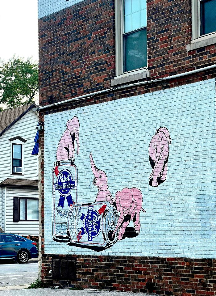 Street art mural showing pink elephants interacting with oversized beer cans on a brick wall in an urban setting.