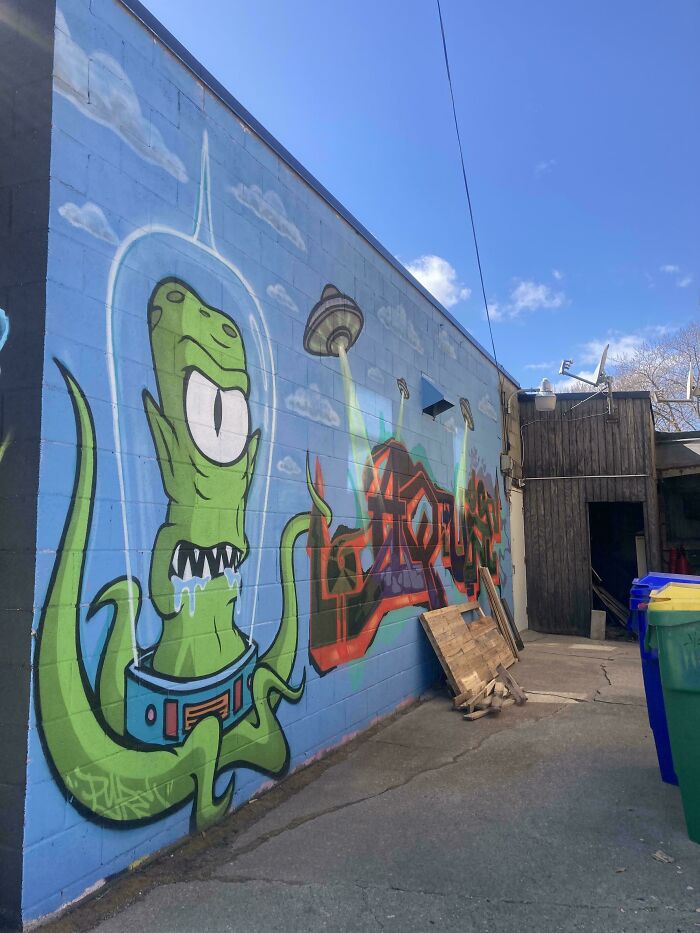 Colorful and powerful street art of a green alien with one eye and a UFO on a blue brick wall outdoors.