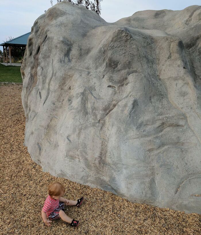 Toddler sitting on wood chips near a large rock formation that looks fake but is 100% real, unphotoshopped pic.