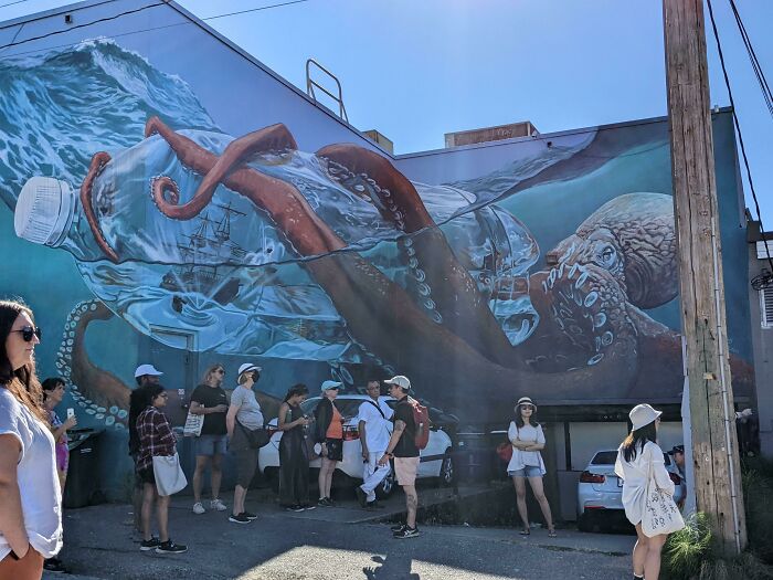 Large street art mural of an octopus inside a plastic bottle with people gathered below, showcasing powerful street art.