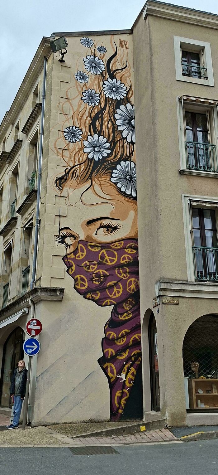 Street art mural of a woman with flowers in her hair and a peace sign-patterned mask on a building corner.