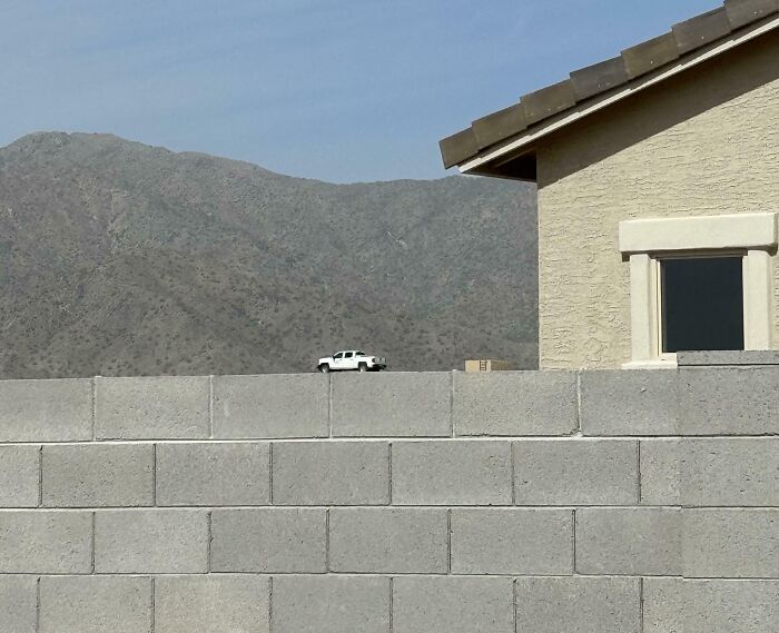 Small white truck appears tiny on a rooftop behind a concrete wall in an unphotoshopped real photo that looks fake.