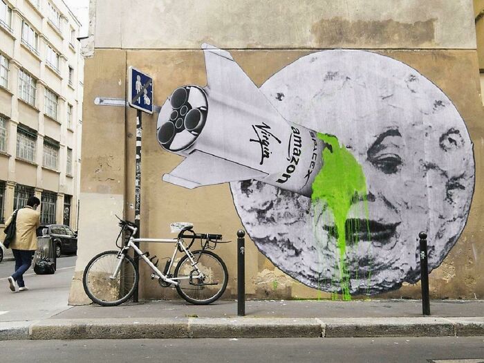 Street art mural of a rocket labeled Amazon and Virgin hitting a face of the moon with green paint on a city wall.