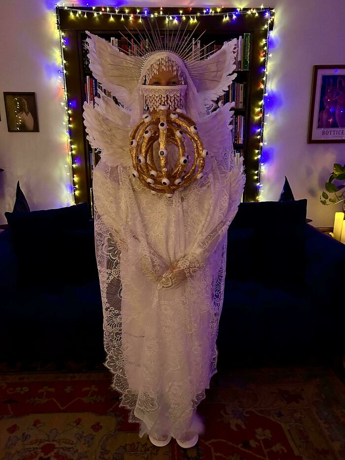 Person wearing a creative Halloween costume with lace, wings, and an eye-covered crown in a dimly lit room.