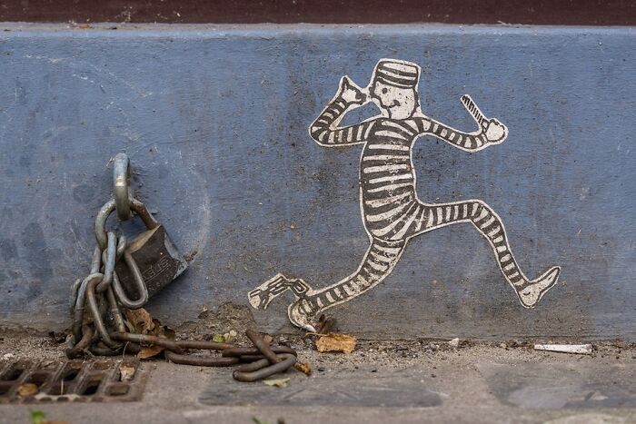 Street art of a running prisoner in striped clothes near a chained padlock on a concrete wall and sidewalk.
