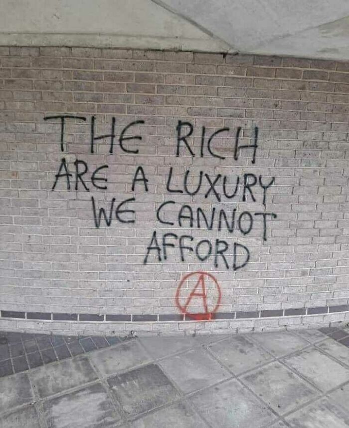 Street art message on brick wall stating the rich are a luxury we cannot afford, with an anarchist symbol below.