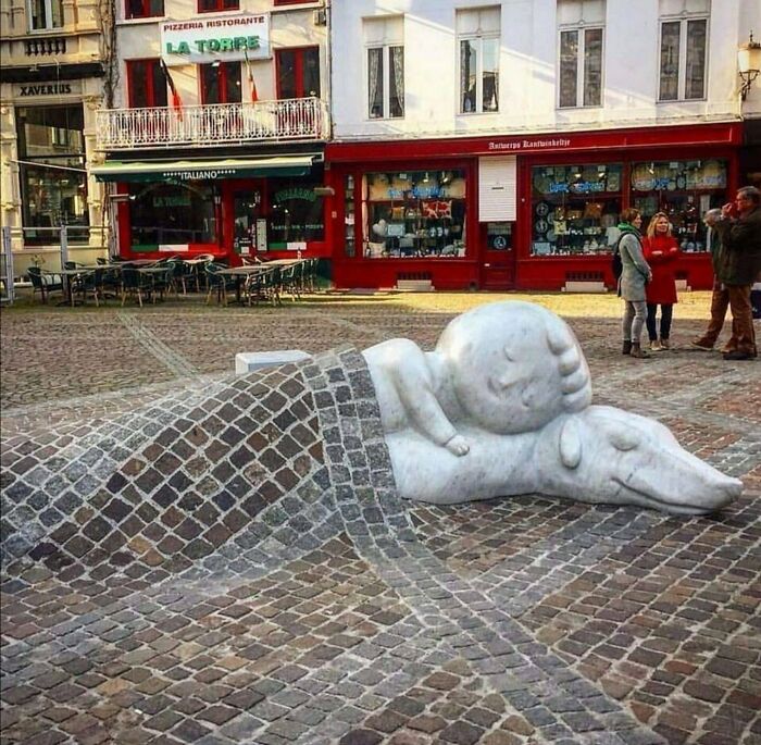Street art sculpture of a child sleeping on a dog, integrated with cobblestone pavement in a public square.