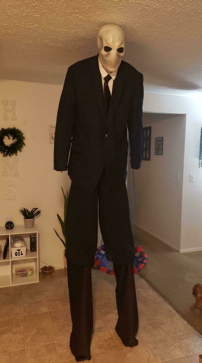 Tall figure in a black suit and eerie mask Halloween costume, standing in a living room for Halloween party inspiration.