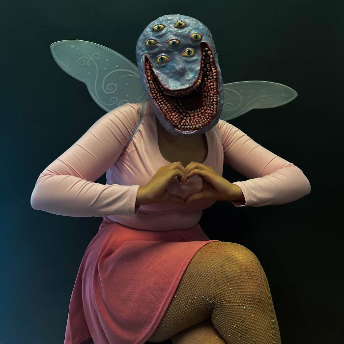 Person in creative Halloween costume with multiple eyes mask and fairy wings posing with hand heart gesture.