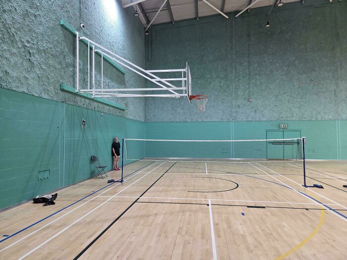 Indoor basketball court with a misaligned basketball hoop blocking the badminton net, mildly infuriating scene testing patience.