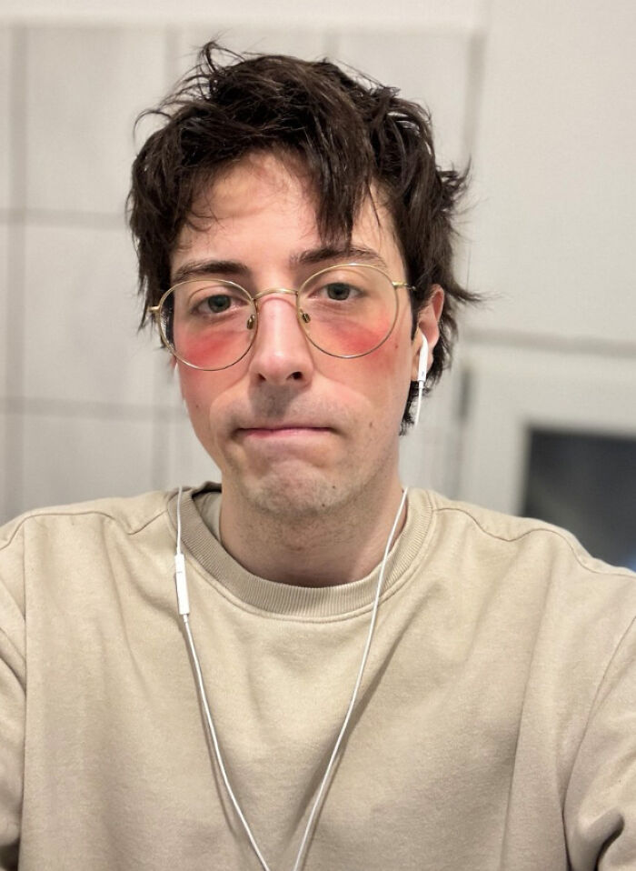 Young man with messy hair and glasses looking mildly infuriated, wearing earbuds and a beige sweatshirt indoors.