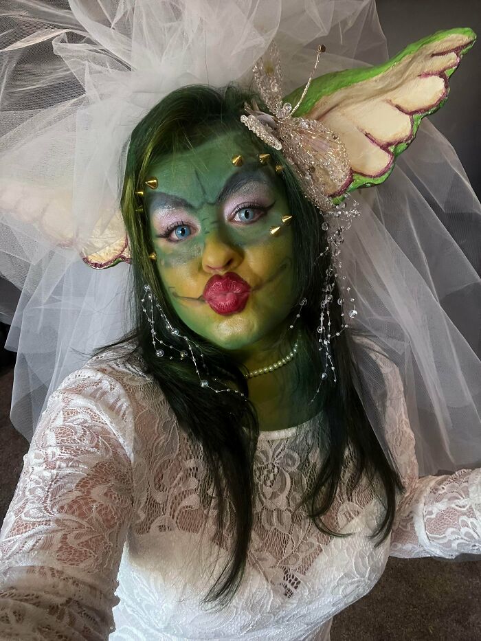 Person in a genius Halloween costume with green face paint, elf ears, spikes, and white lace dress posing for a selfie.