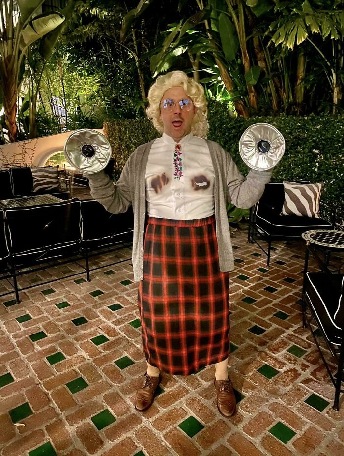 Person in a creative Halloween costume with a blonde wig, cardigan, and plaid skirt holding makeshift cymbals outdoors.
