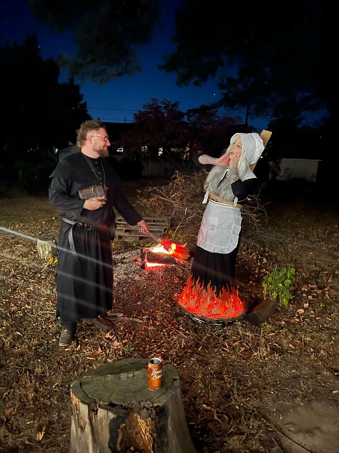 Man dressed as a priest playfully roasting a woman in a Halloween costume with flames on her skirt by a fire pit.