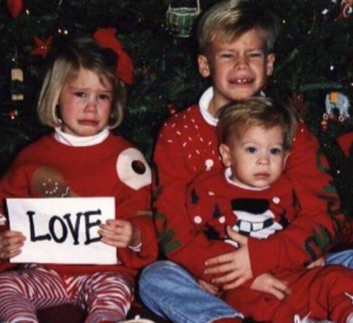 Three children in holiday sweaters sitting by a Christmas tree with awkward and cringed family photo expressions.