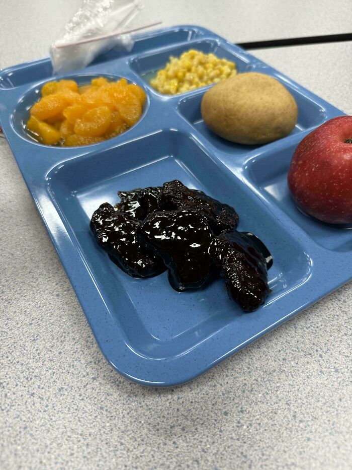 Blue cafeteria tray with unappetizing food including glossy black meat, canned fruit, corn, a bread roll, and an apple.