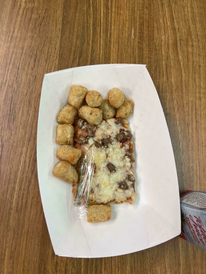 Unholy food pic of a small pizza slice with irregular tater tots in a paper tray on a wooden table.