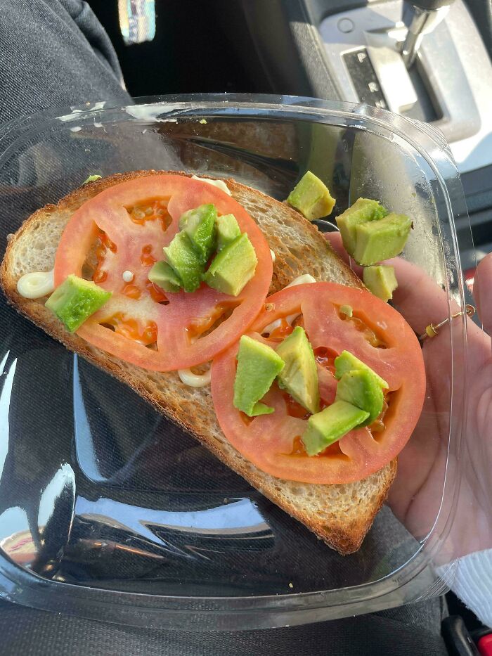 Unholy food pic of open-faced sandwich with tomato slices and diced avocado on toasted bread in plastic container.