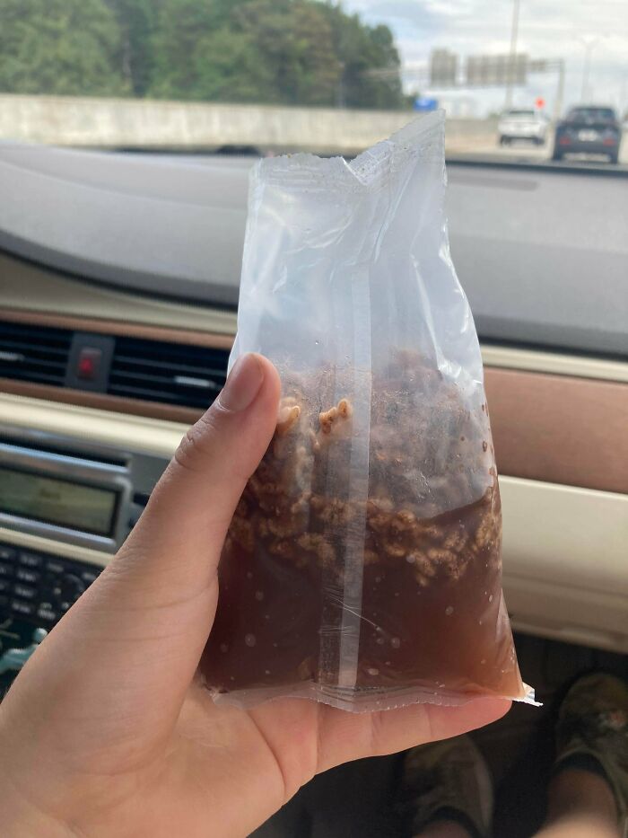Hand holding a sealed plastic bag containing unappetizing food with an unholy texture inside a car.