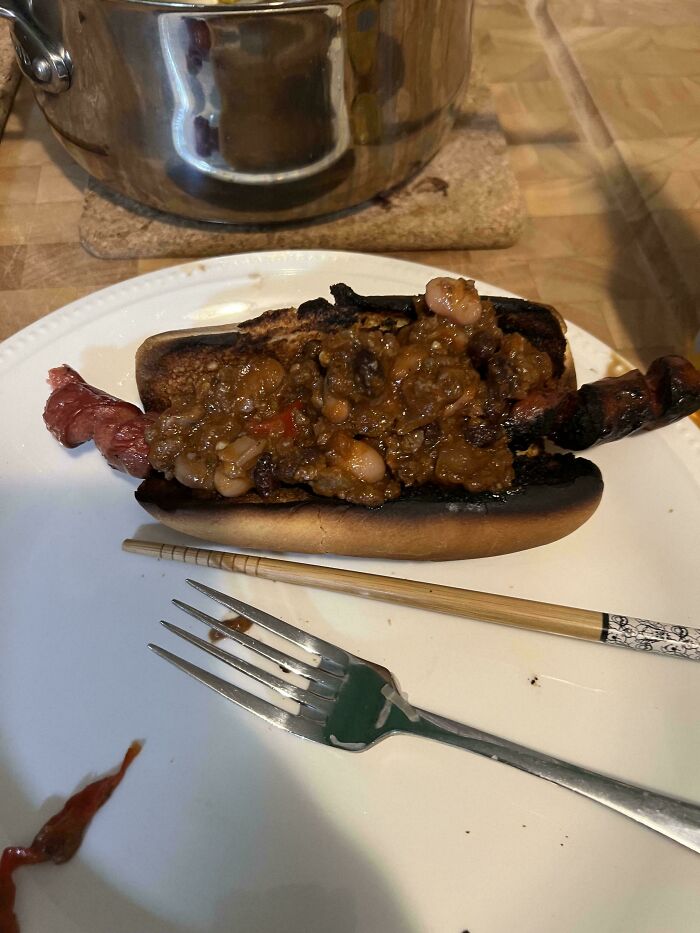Burnt hot dog with unappetizing chili and beans on a white plate, showing unholy food pics that look anything but appetizing.