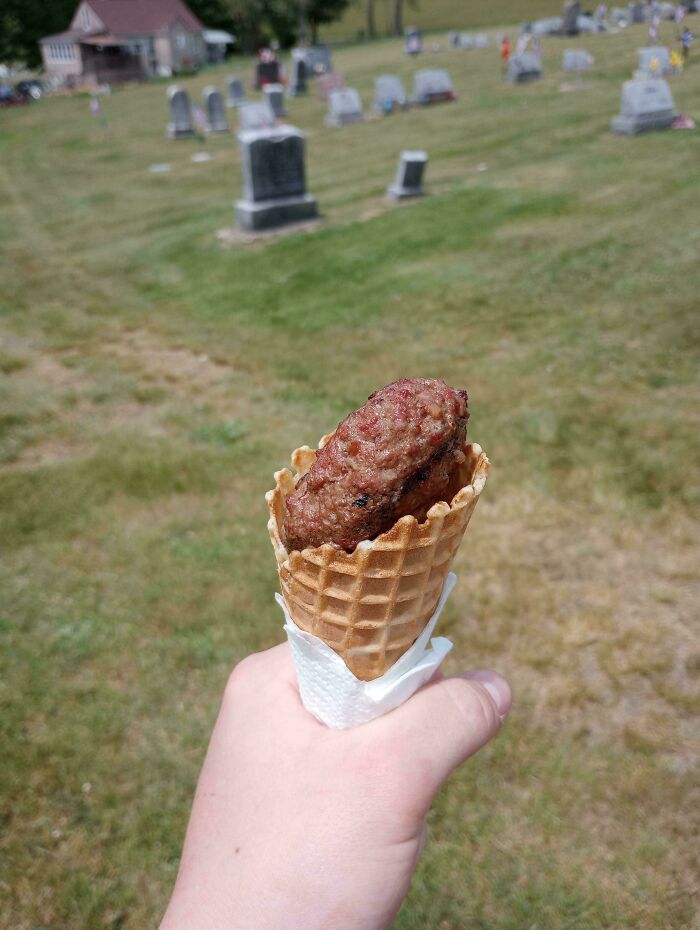 Hand holding a waffle cone filled with a cooked meat patty, unholy food pics that look anything but appetizing in a cemetery.
