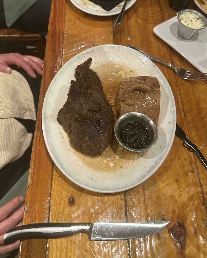 Plate with unappetizing food showing a dark, overcooked piece of meat and a bread roll with sauce on a wooden table.