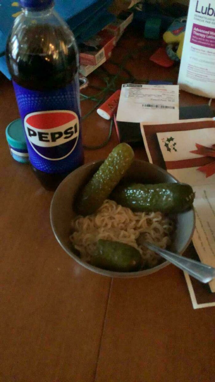 Bowl of instant noodles topped with whole pickles next to a bottle of Pepsi on a cluttered wooden table unholy food pics.