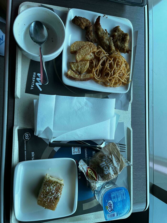 Hospital tray with unappetizing fried food, potato wedges, spaghetti, bread, and packaged water and butter.