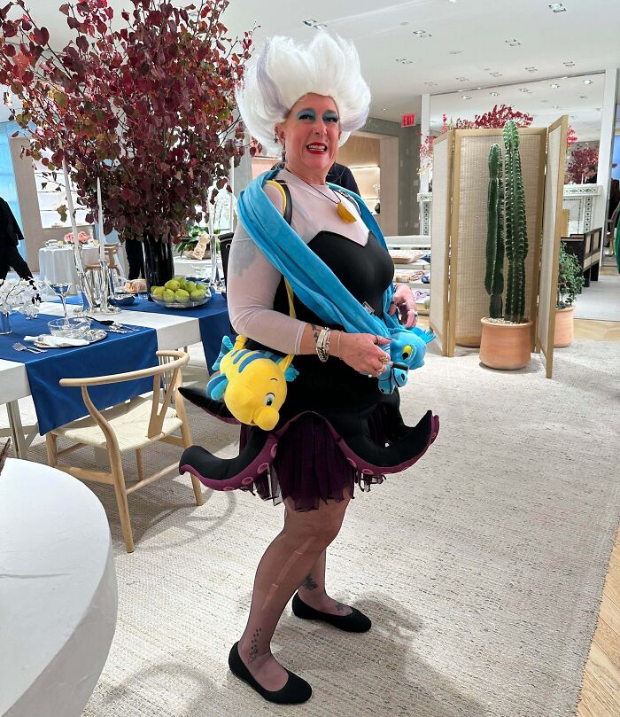 Woman in a genius Halloween costume inspired by Ursula, wearing an octopus skirt and carrying Flounder plush toys indoors.